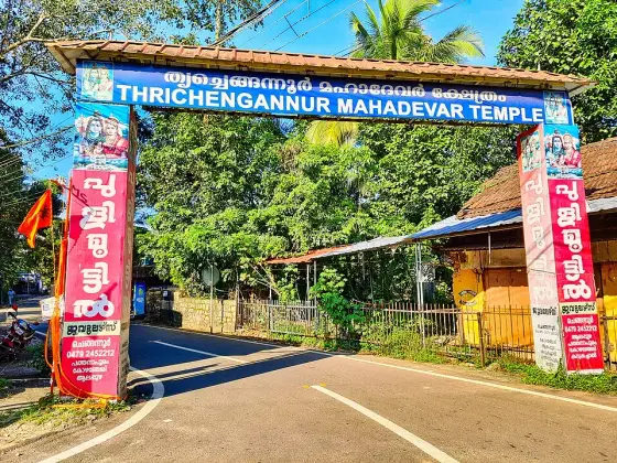 The roadside arch. Its board carries the temple’s own name-form — Thrichengannur Mahadevar Temple — which is why it is shown; the red panels down both piers are a jewellery firm’s sponsorship, not part of the temple.