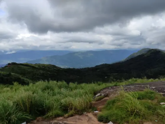 Dark clouds hang over layered hills beyond grass and exposed rock at Ilaveezhapoonchira.