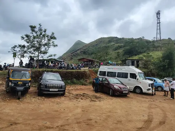 Cars, a minibus and an autorickshaw stand on Illikkal Kallu’s earth parking area, below a terrace of motorcycles and small shelters.