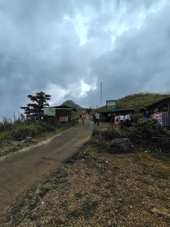 Visitors descend the paved approach past small stalls and the green Illikkal Kallu tourism sign, with the hill rising behind.