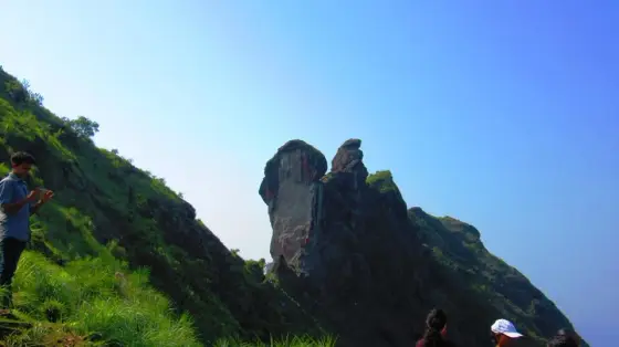 Visitors stand on a grassy slope below Illikkal Kallu’s rock formations in this older photograph; the pictured positions do not establish today’s permitted walking route.