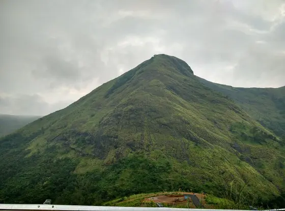 A broad grass-covered hill rises beyond a guardrail and a lower bend of the Illikkal Kallu approach.