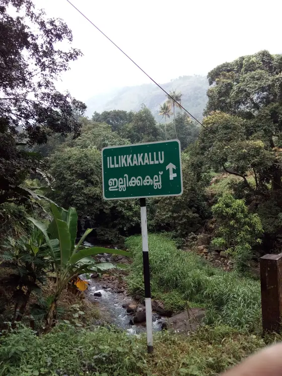 A green sign reading ILLIKKAKALLU in English and Malayalam points ahead above a rocky stream and green vegetation.