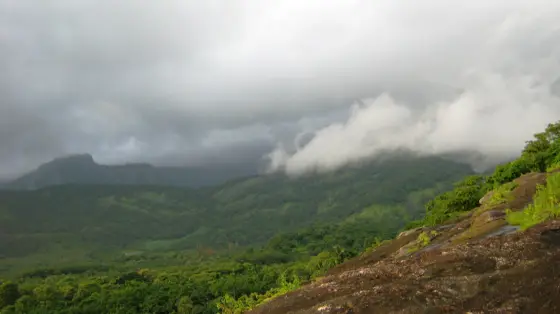 Low cloud crosses wooded Dhoni hills beyond a broad sloping rock surface.
