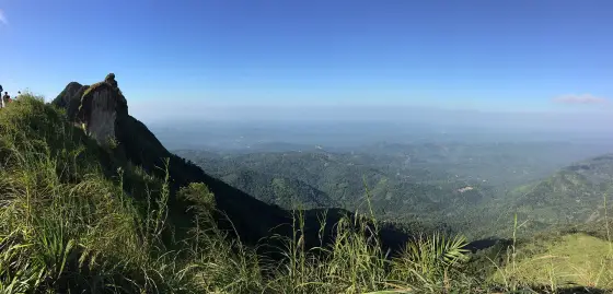 Illikkal Kallu stands at the left of a wide view across wooded valleys beneath a blue sky.
