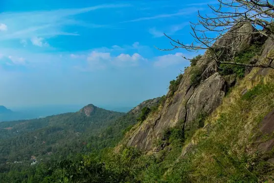 A steep exposed rock slope at Kurumbalakotta overlooks wooded hills beneath a blue sky.
