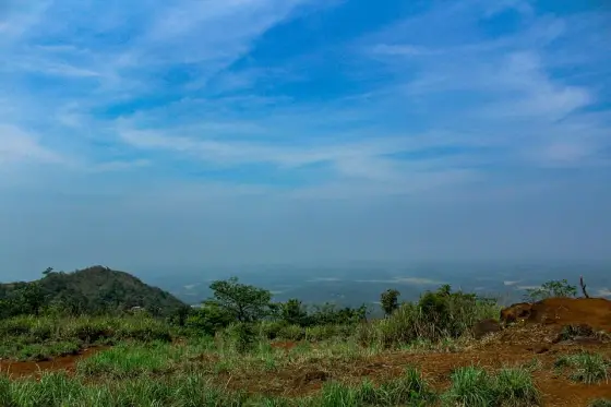 Grass and red earth on Kurumbalakotta overlook a lower hill and a hazy distant landscape.