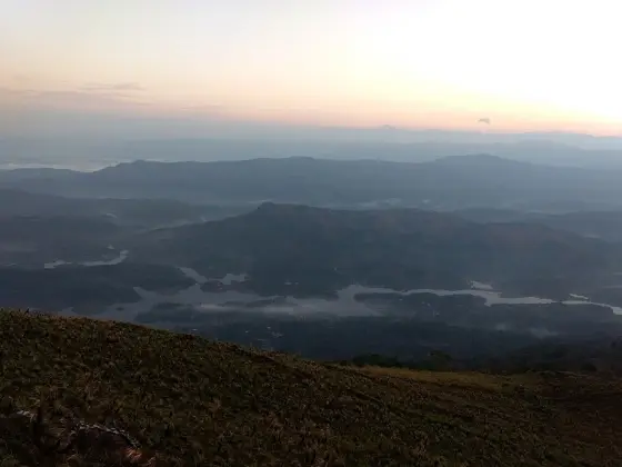 Layers of dark hills and branching water lie below Ilaveezhapoonchira beneath a pale pink and yellow sky.
