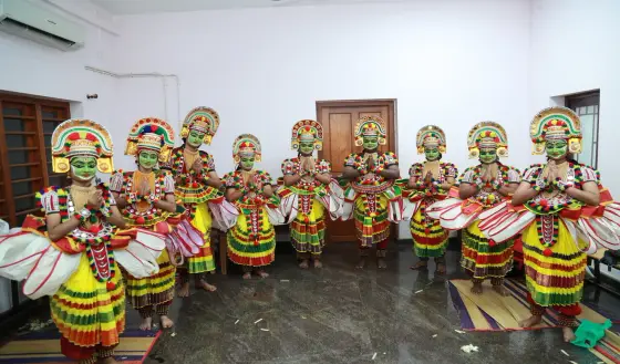 Performers with green-painted faces, decorated headdresses and striped full skirts stand together inside a Kalamandalam room.