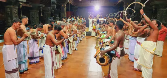 Two lines of musicians play drums, cymbals and curved horns beneath carved pillars at Kerala Kalamandalam.
