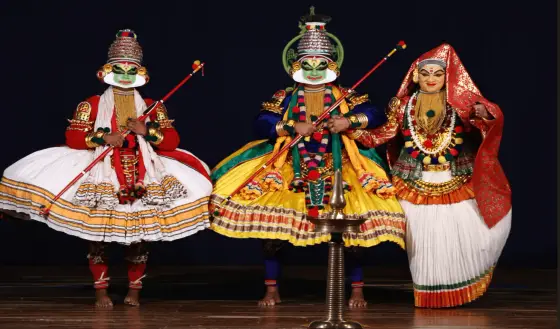 Three costumed performers stand beside a stage lamp in a photograph from Kalamandalam’s performing-arts gallery.