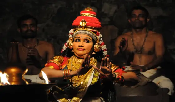 A performer holds both hands in a gesture beside a lamp, with two seated accompanists behind her, in Kalamandalam’s Nangyarkooth photograph.