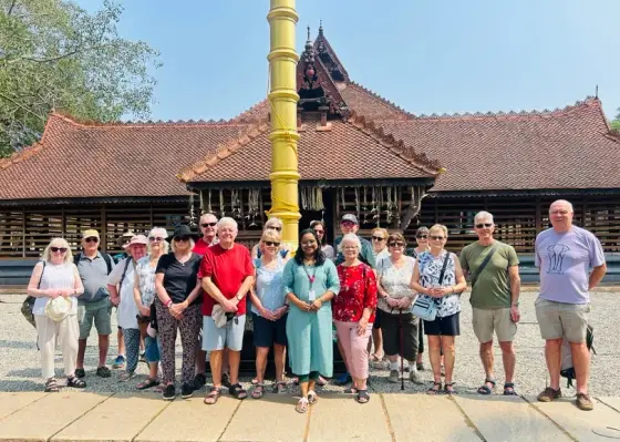 A visiting group stands beside the golden flagstaff in front of Kalamandalam’s timber-sided Koothambalam.