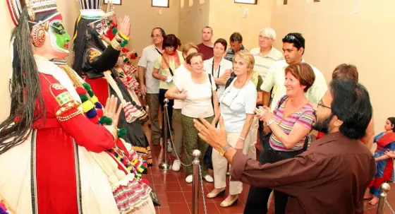 A guide explains costumed Kathakali figures to visitors standing behind a low chain barrier in the art gallery.