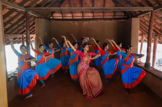 A teacher and dance students practise raised-arm gestures beneath the exposed timber roof of a Kalamandalam teaching room.