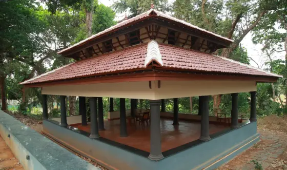 An open-sided teaching pavilion has dark columns, a red floor and a tiered tiled roof among trees.