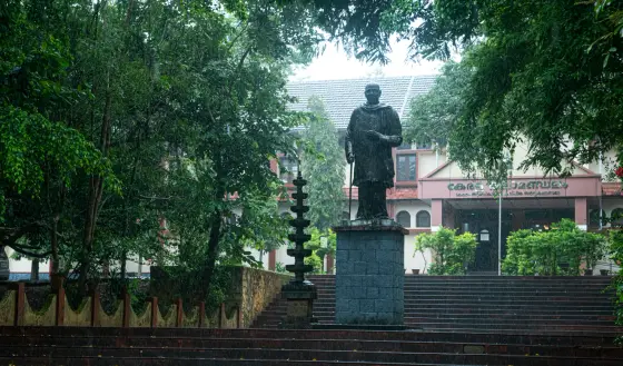 A statue holding a walking stick stands among trees before the main Kalamandalam building, with wet steps and a tall lamp nearby.