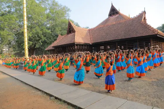 Rows of dance students in blue and green with orange sashes practise outside the tiled-roof Koothambalam.