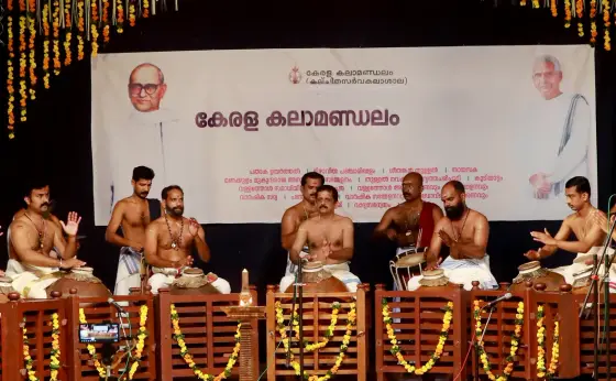 Seated musicians play pot-shaped Mizhavu drums behind a flower-decorated rail at a Kerala Kalamandalam event.