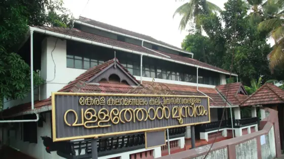 A Malayalam sign for the Vallathol memorial museum stands in front of a white building with tiled roofs and screened upper windows.