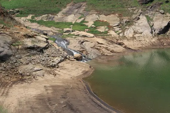 A low cascade runs across bare rock towards green reservoir water at Anchuruli.