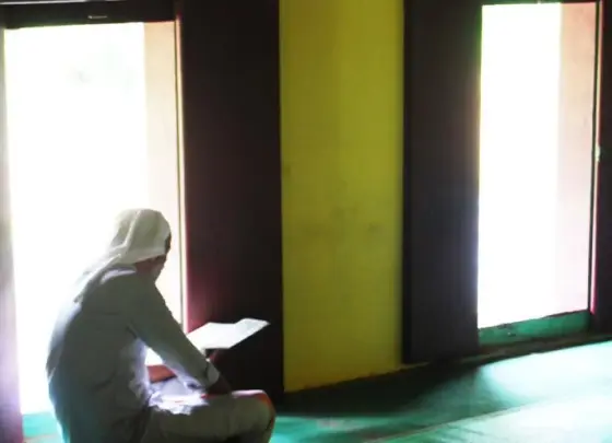 A reader sits beside the maqaam’s bright doorway and dark timber interior.