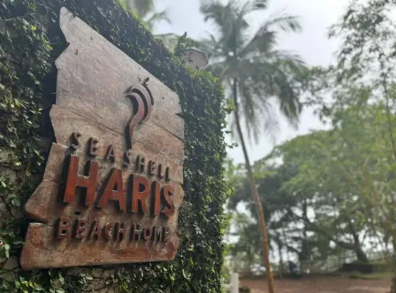 A wooden Seashell Haris Beach Home sign stands against ivy, with palms behind.