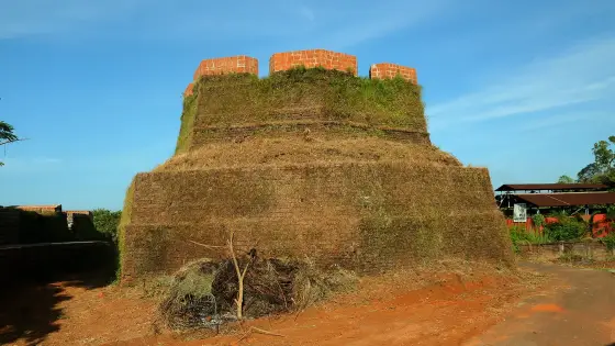 A frontal view shows the rounded two-tier bastion, red upper projections and dry vegetation at Hosdurg.