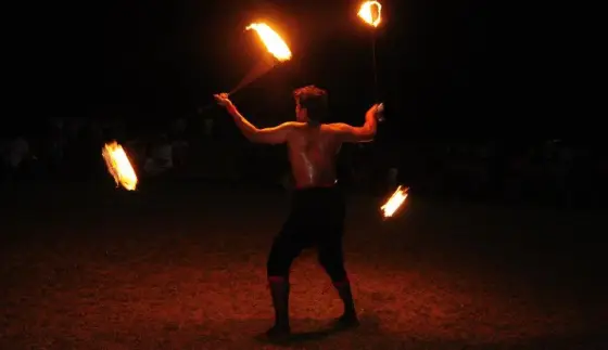 An archival festival performer handles burning props against the night sky.