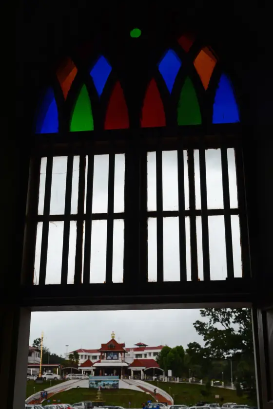 Coloured window panes frame the neighbouring Alphonsa pilgrimage buildings; this is a shrine-precinct view, not the parish sanctuary.