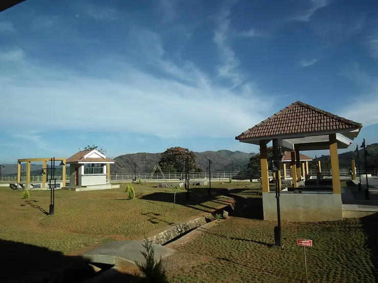 A tiled pavilion, a small white ticket hut and swing frames on the mown upper terrace of Hill View Park, with high ridges behind