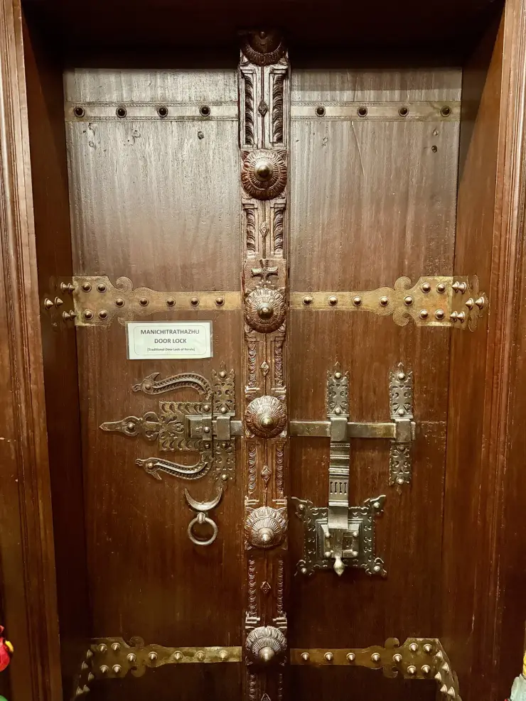 A carved teak door at the Kerala Folklore Museum fitted with an ornate brass manichitrathazhu lock, bolts and studded straps