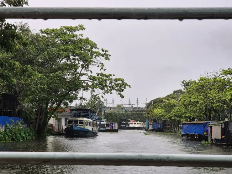 A wide canal basin at Alappuzha in heavy rain, ferry boats moored along both banks and a footbridge crossing beyond