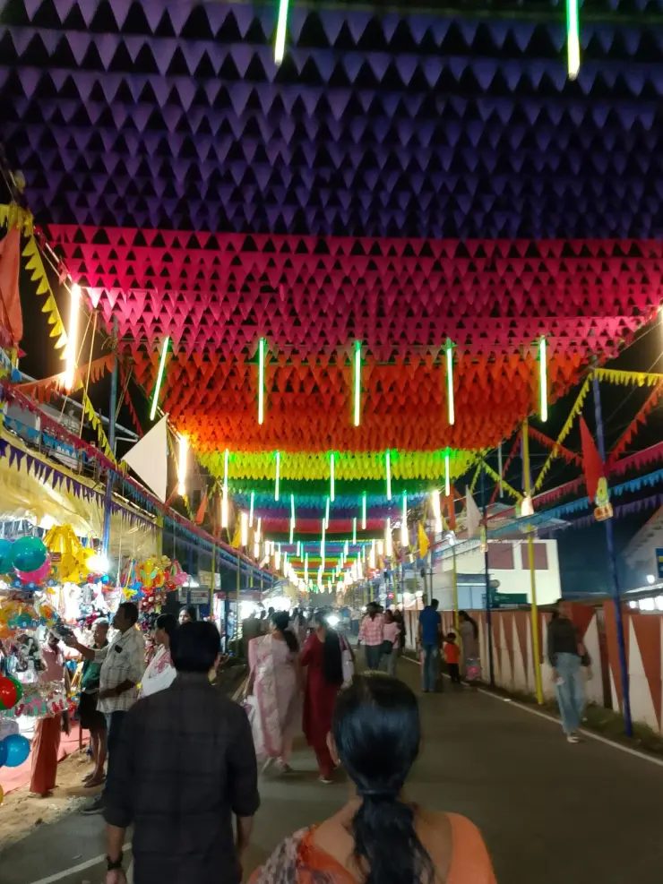 A street in Alappuzha roofed over with brightly coloured pleated decorations and strings of lights during Mullakkal Chirappu, crowded with people and stalls