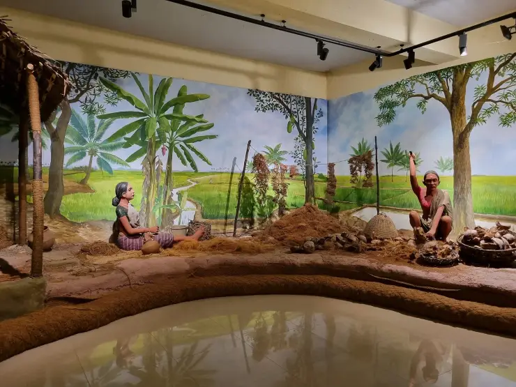 A walk-in museum diorama of coir retting, with life-size painted figures beside a backwater channel and a reflecting pool set into the floor