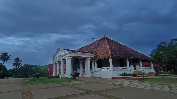 Kanakakunnu Palace at dusk under a heavy sky, its white classical portico set against a tiled hipped roof and red walls
