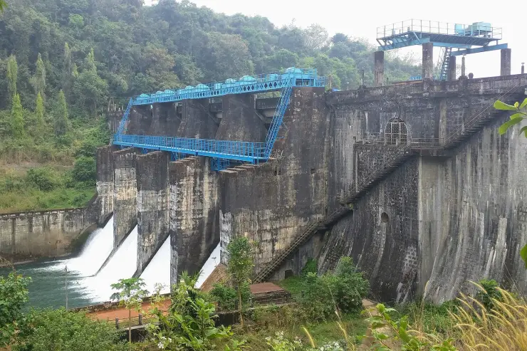 The stone masonry gravity dam at Peruvannamuzhi seen from the abutment, with a blue gantry crane over the spillway bays and water running down the ogee face.
