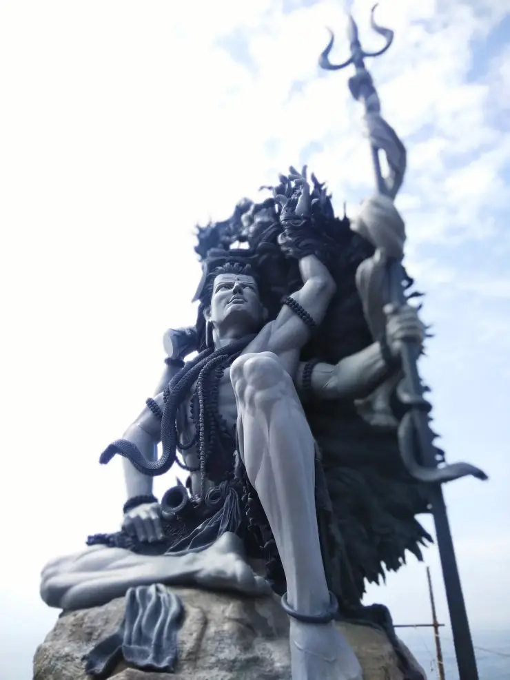 Low-angle view of the Aazhimala Gangadhareswara sculpture against a bright sky, with the raised trishul at the right