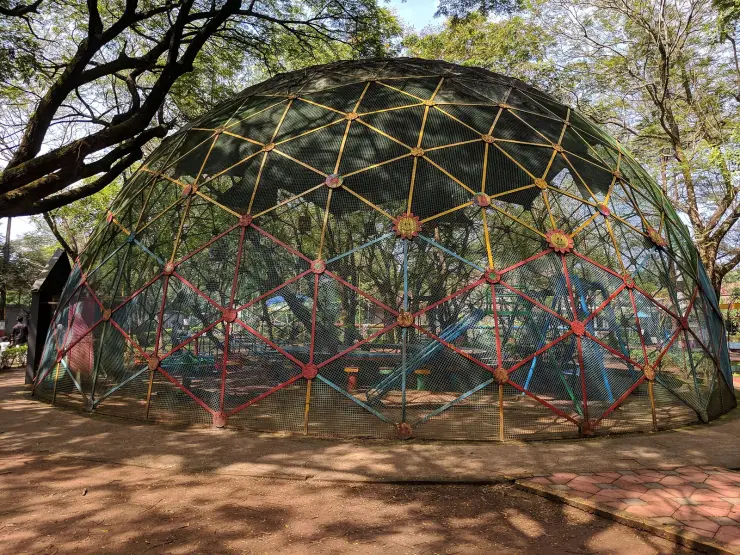 A large multicoloured geodesic dome of red, blue and yellow struts covered in dark mesh, standing over a play area under mature shade trees in the Science Park at Kozhikode.