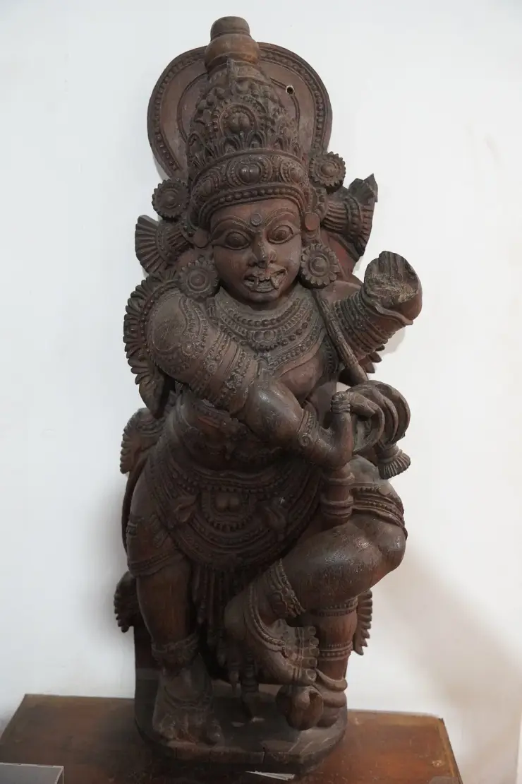 A carved wooden door guardian, crowned and wide-eyed with tusked mouth, heavily ornamented, standing in a museum gallery against a white wall