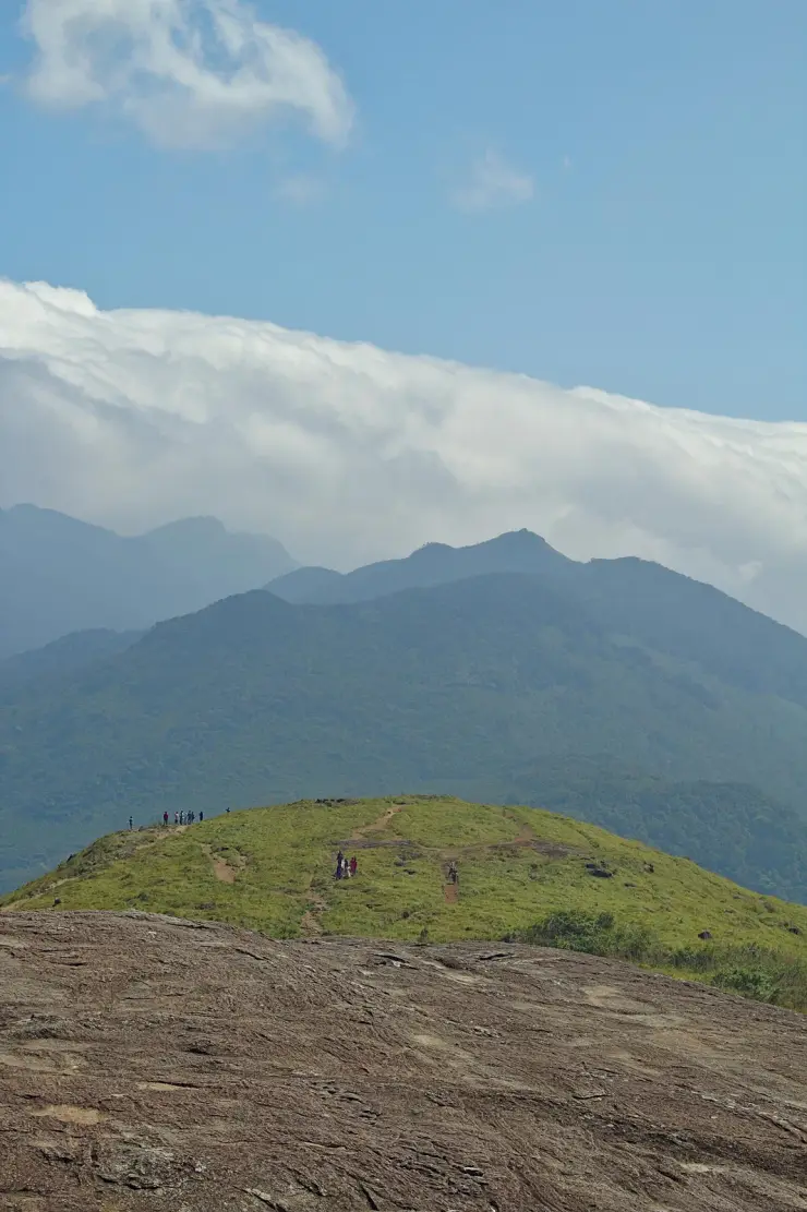 A green grass-topped shoulder with tiny distant figures, forested ridges behind it and a thick bank of cloud spilling over the skyline