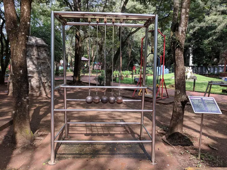 A full-size Newton’s cradle in polished steel in the Science Park at Kozhikode: five iron spheres hanging in a tall tubular frame on a paved pad under mature shade trees, with an explanatory plate beside it.