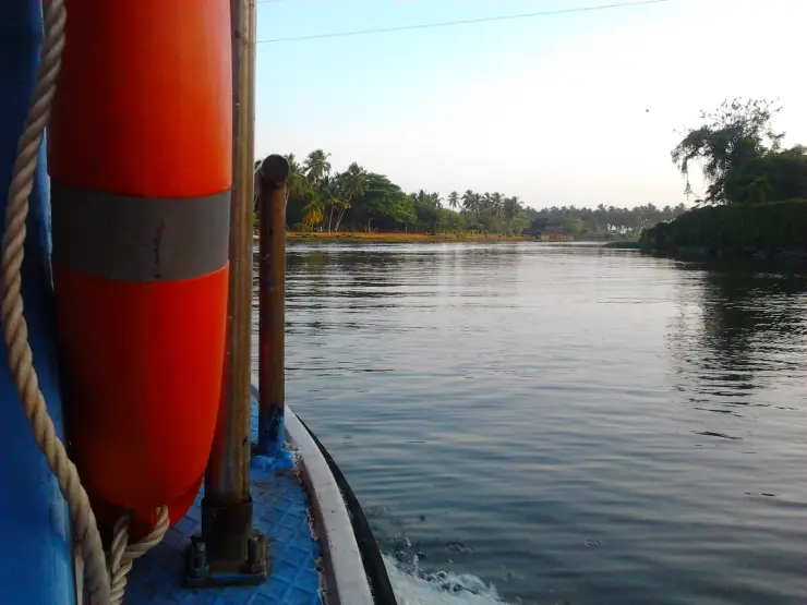 The bow of a boat with an orange life buoy, running on the Veli backwater past wooded banks