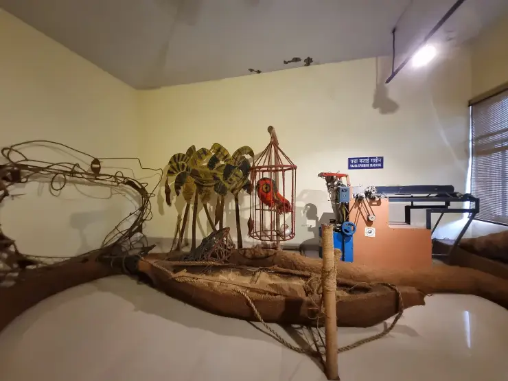 A museum gallery with a labelled Vajra spinning machine at the right, a full-size coir-clad country boat across the foreground and a coir fishing net at left