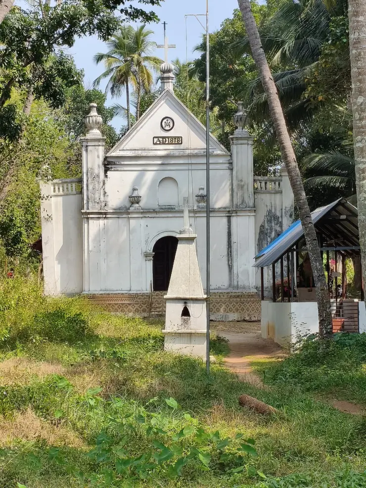 The white-plastered west front of the Dutch Church at Munroe Island: a gabled pediment carrying a monogram in a circular roundel above a carved plaque reading A.D. 1878, urn finials at the corners, a cross at the apex, a round-arched central doorway with a dark timber door, and a band of patterned glazed tiles along the base. Uncut grass in the foreground.