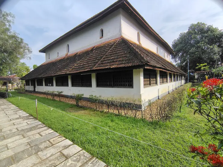 The tiled two-storey Koyikkal Palace seen from its garden path, with a long run of dark latticed windows under deep eaves