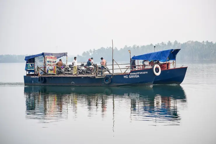 A blue-hulled flat-decked ferry named MC Ganga crossing calm grey-green water on Ashtamudi, about a dozen motorcycles parked across its open deck under a blue tarpaulin canopy, a few passengers standing among them and a low palm-covered shore in the haze behind.