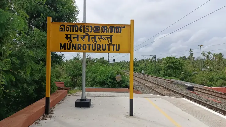 The yellow station nameboard at Munroturuttu reading the name in Malayalam and Roman script, on a freshly concreted platform with a yellow safety line, looking down a double electrified track between coconut palms under an overcast sky.
