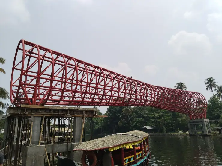 A red steel lattice footbridge over water at Punnamada, tapering from a wide flared mouth to a narrow tail like a snake boat