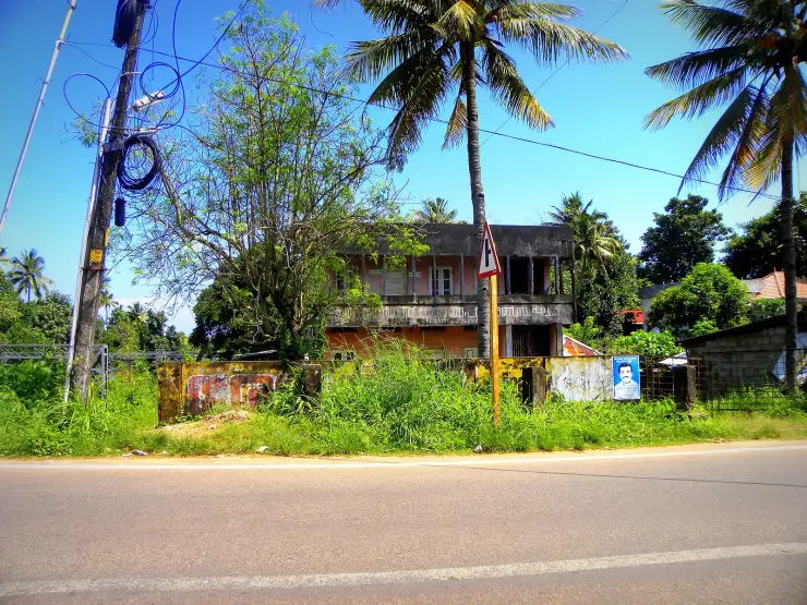 The Paravur Coir Industries Co-operative Union office seen from across a road: a two-storey concrete building, weather-stained and streaked with damp, its balcony and windows dark and empty, half swallowed by tall grass and scrub, with coconut palms behind and a Malayalam signboard barely legible on the façade.